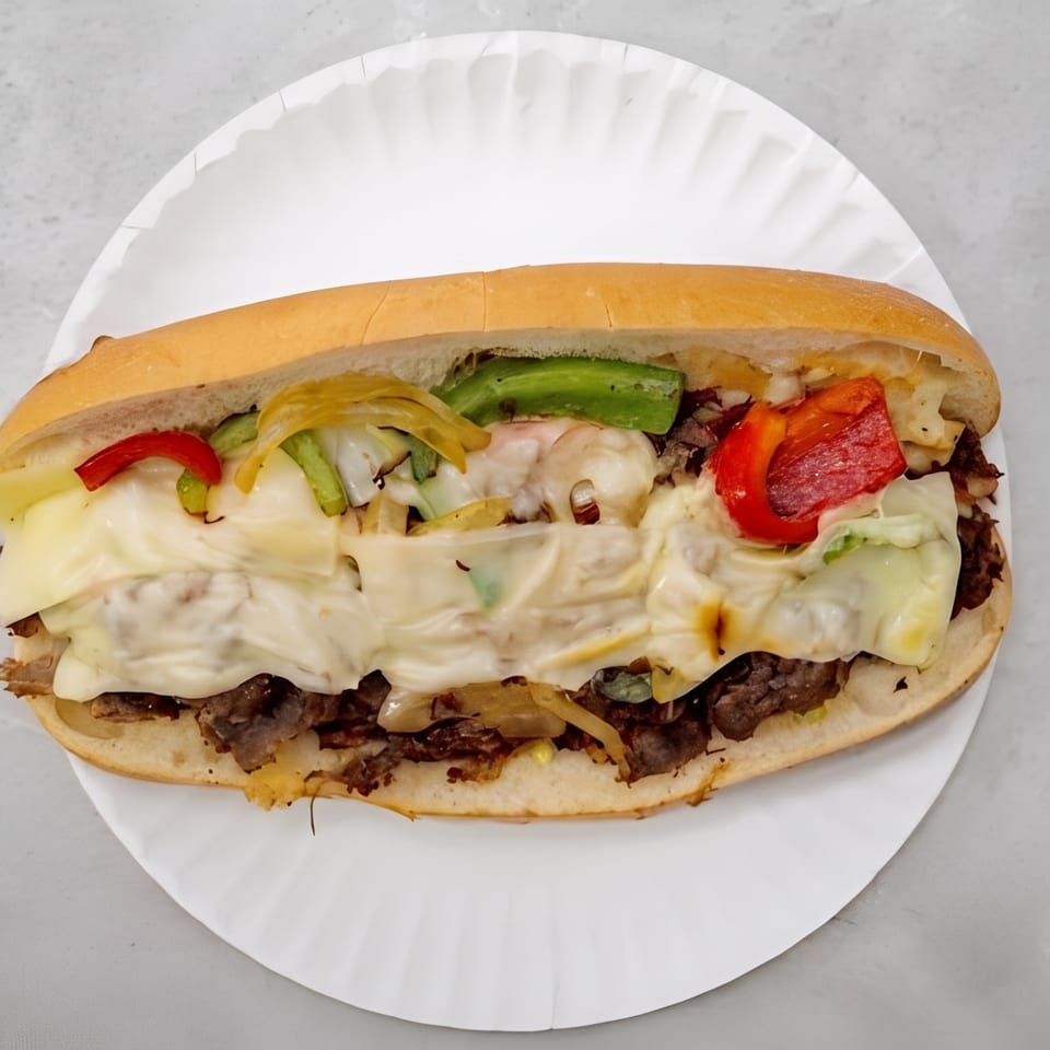 Hearty cheesesteak sandwich with melted cheese, sliced beef, grilled peppers, and onions on a white paper plate.