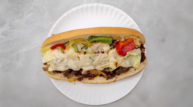Hearty cheesesteak sandwich with melted cheese, sliced beef, grilled peppers, and onions on a white paper plate.