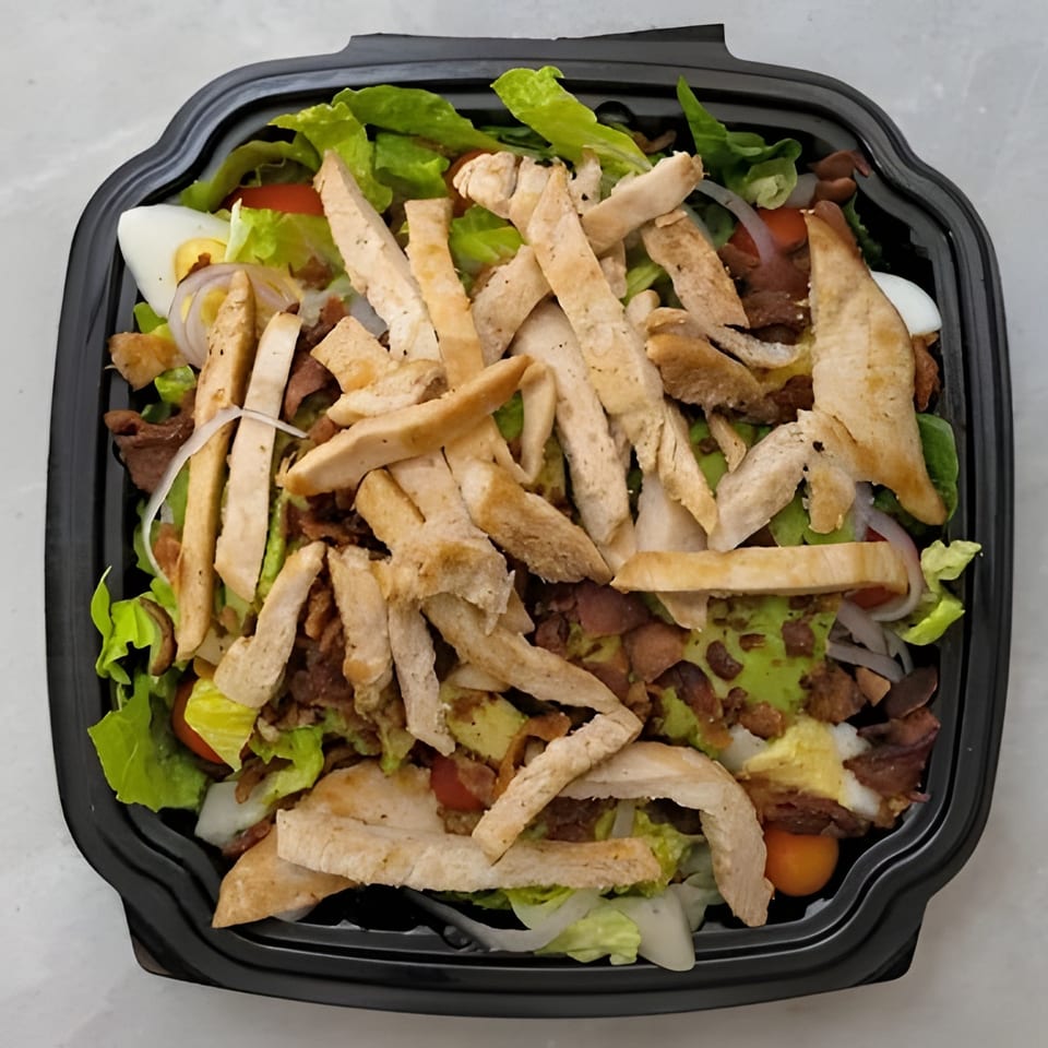 Chicken salad with lettuce, bacon, avocado, egg, and tomatoes in a black takeout container.