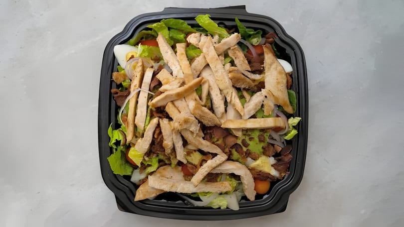 Chicken salad with lettuce, bacon, avocado, egg, and tomatoes in a black takeout container.